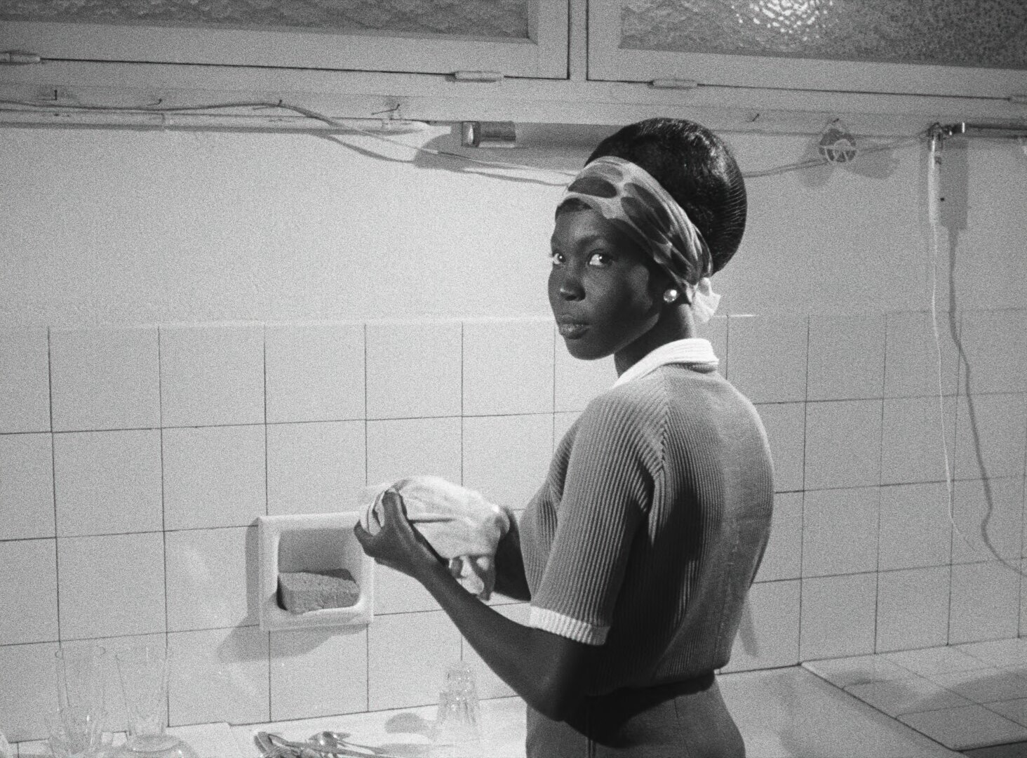 Diouana washes dishes in a tiled kitchen, glancing over her shoulder at the camera — a still from Black Girl (La Noire de..., 1966) by Ousmane Sembène