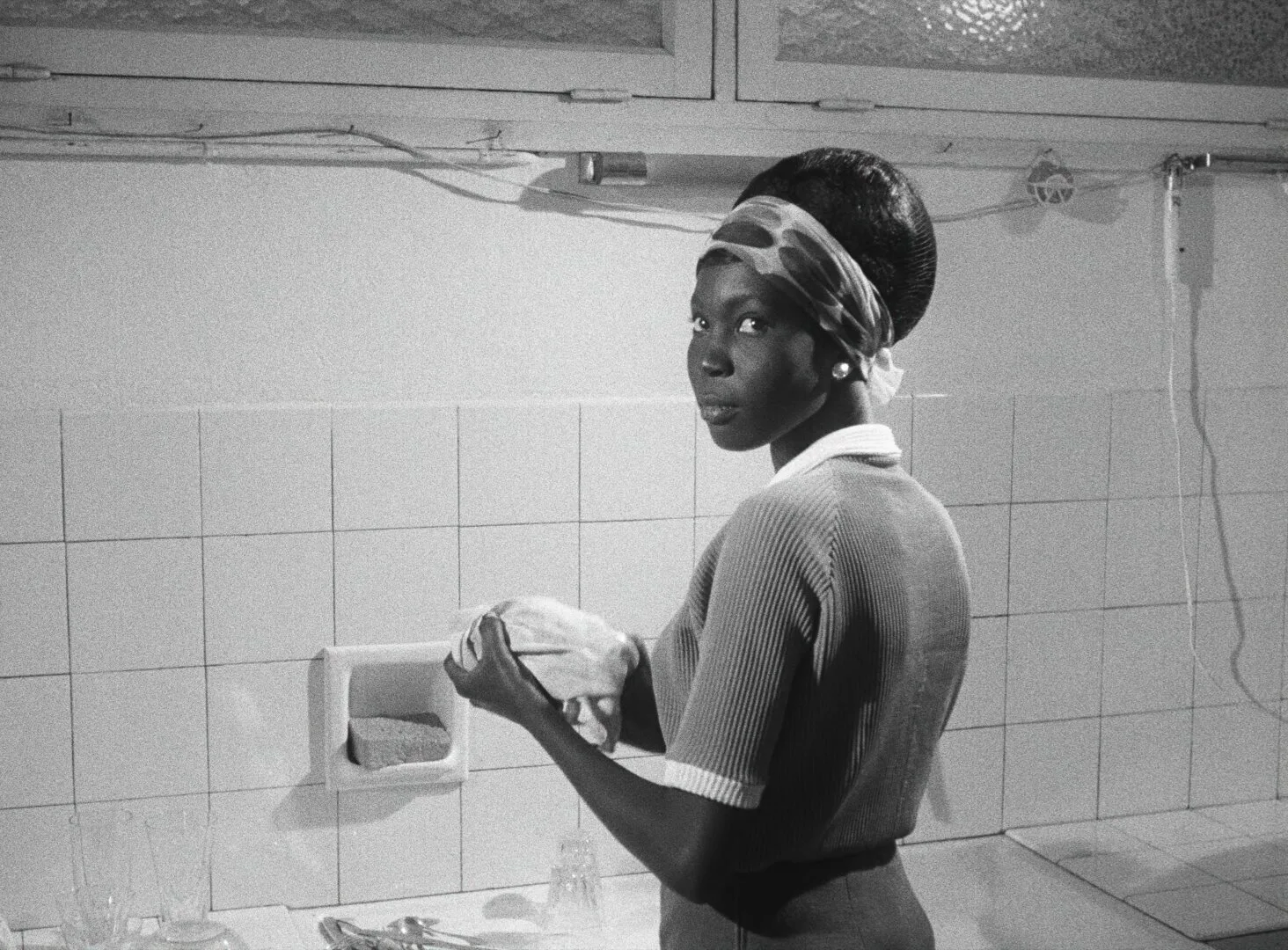 Diouana washes dishes in a tiled kitchen, glancing over her shoulder at the camera — a still from Black Girl (La Noire de..., 1966) by Ousmane Sembène