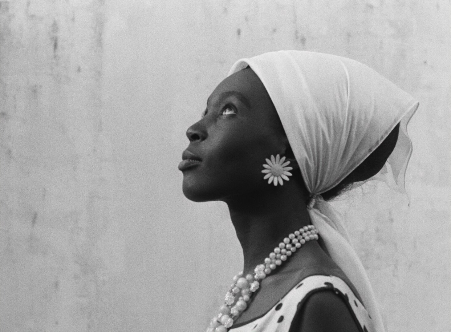 Diouana in profile, gazing upward against a pale wall, wearing a white headscarf and daisy earring — a still from Black Girl (La Noire de..., 1966) by Ousmane Sembène