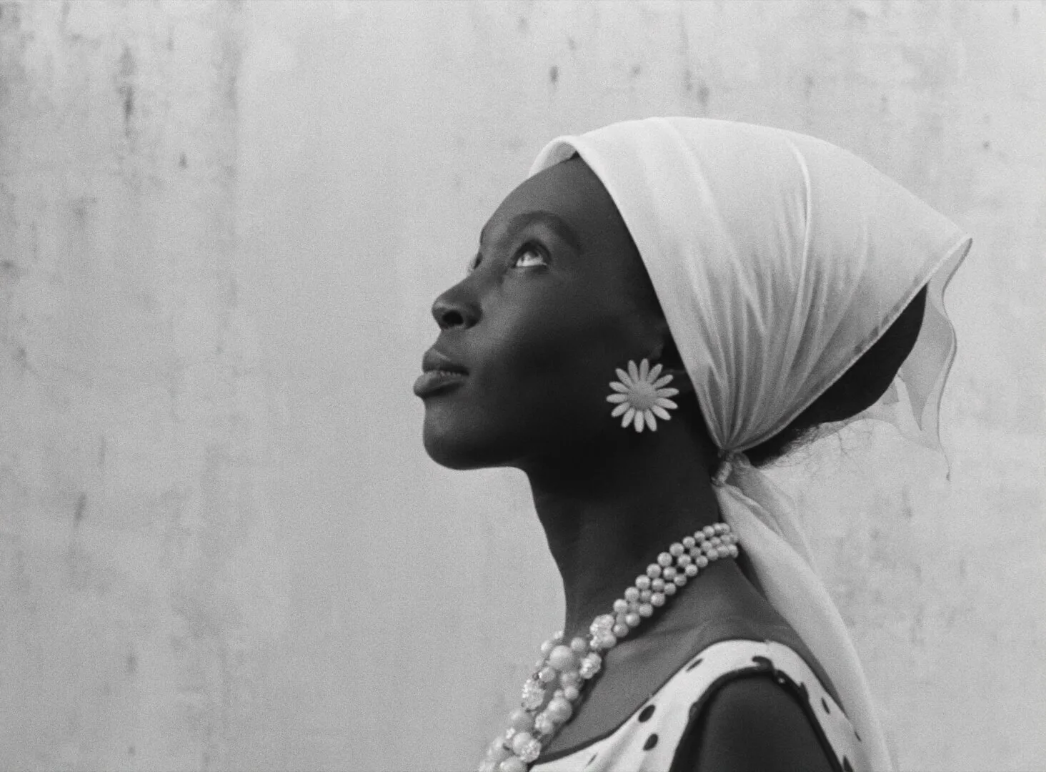 Diouana in profile, gazing upward against a pale wall, wearing a white headscarf and daisy earring — a still from Black Girl (La Noire de..., 1966) by Ousmane Sembène