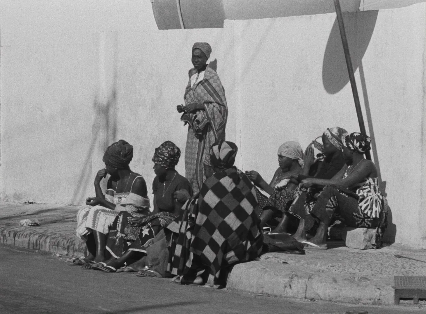 A group of women in traditional dress sit and stand along a whitewashed wall in the Dakar sun — a still from Black Girl (La Noire de..., 1966) by Ousmane Sembène