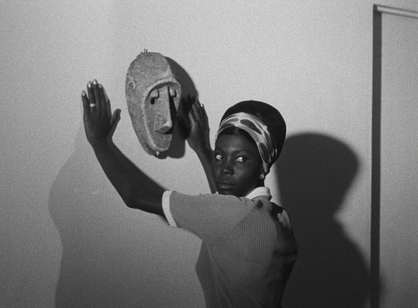 Diouana lifts a carved African mask toward the wall, her shadow spreading behind her, her eyes turned to the camera — a still from Black Girl (La Noire de..., 1966) by Ousmane Sembène