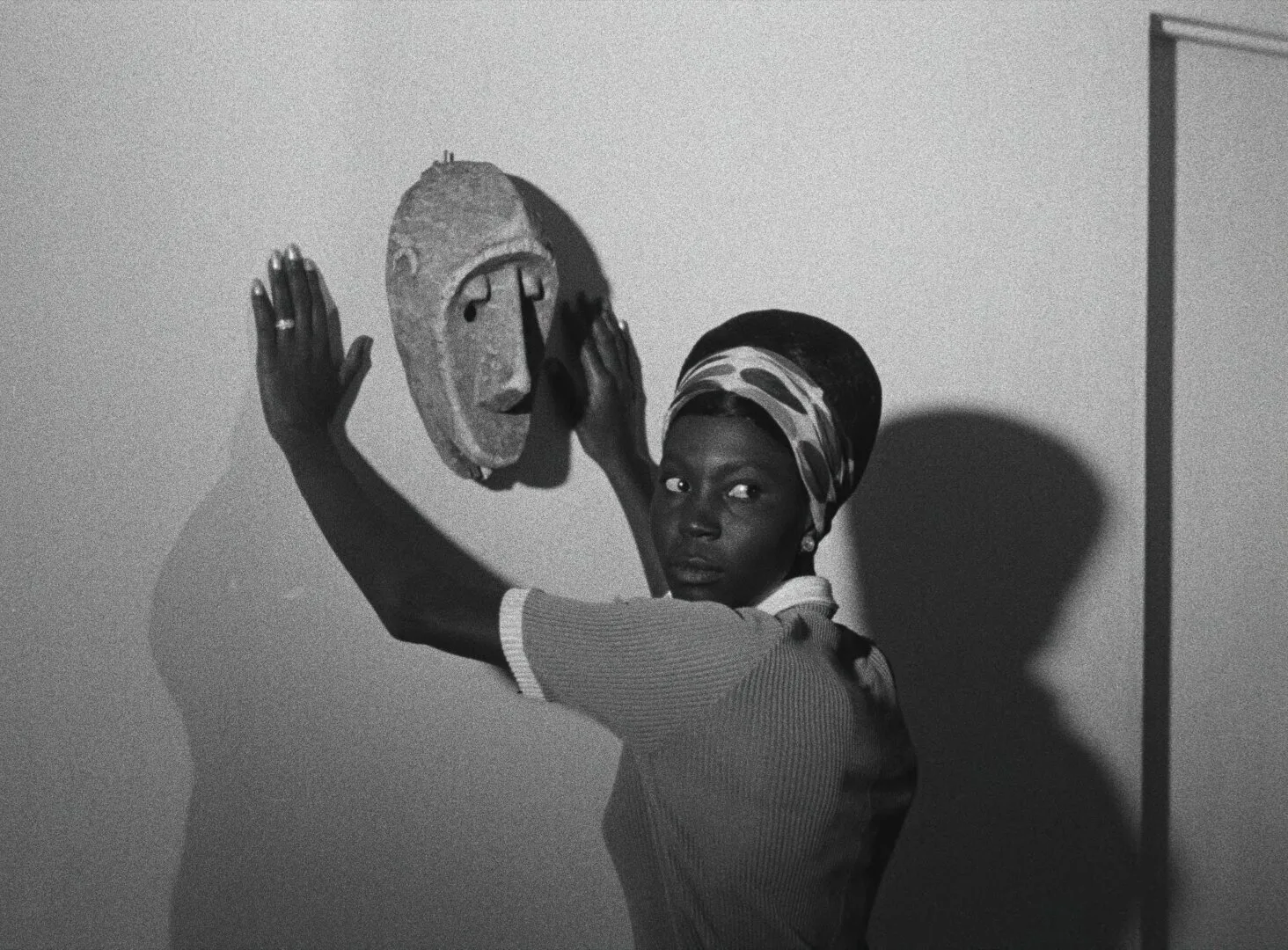 Diouana lifts a carved African mask toward the wall, her shadow spreading behind her, her eyes turned to the camera — a still from Black Girl (La Noire de..., 1966) by Ousmane Sembène