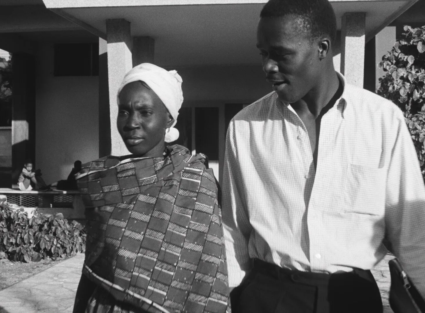 Diouana walks alongside a young man outside a modern building in Dakar, her expression thoughtful — a still from Black Girl (La Noire de..., 1966) by Ousmane Sembène