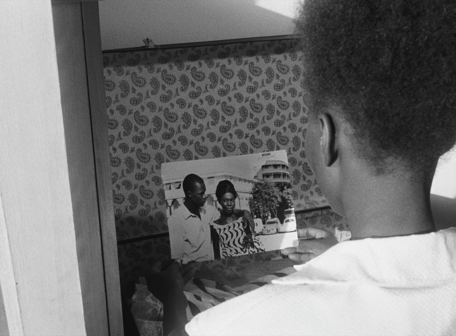 A young man holds a photograph of a couple, viewed from behind in a small room with patterned wallpaper — a still from Black Girl (La Noire de..., 1966) by Ousmane Sembène