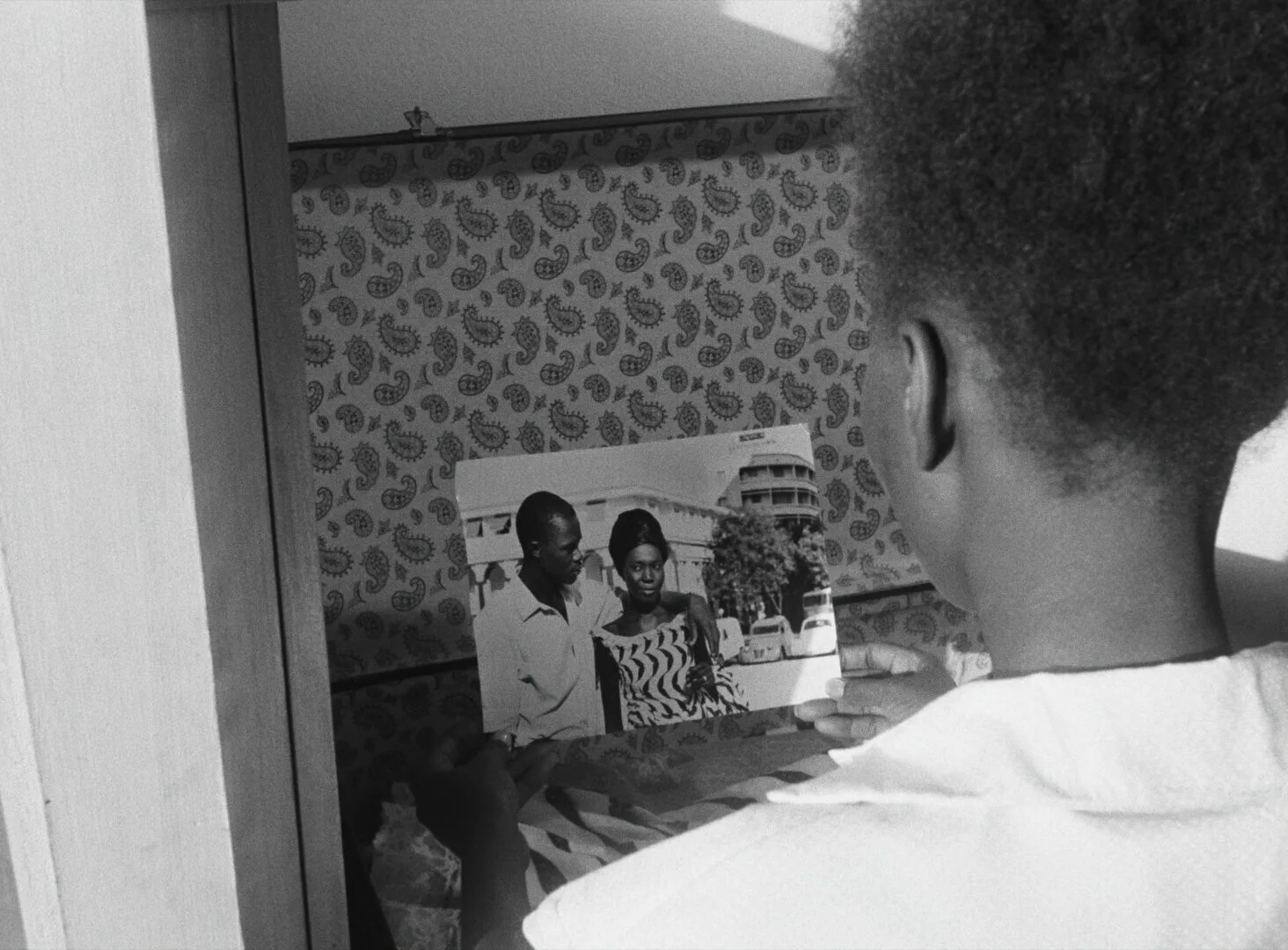 A young man holds a photograph of a couple, viewed from behind in a small room with patterned wallpaper — a still from Black Girl (La Noire de..., 1966) by Ousmane Sembène