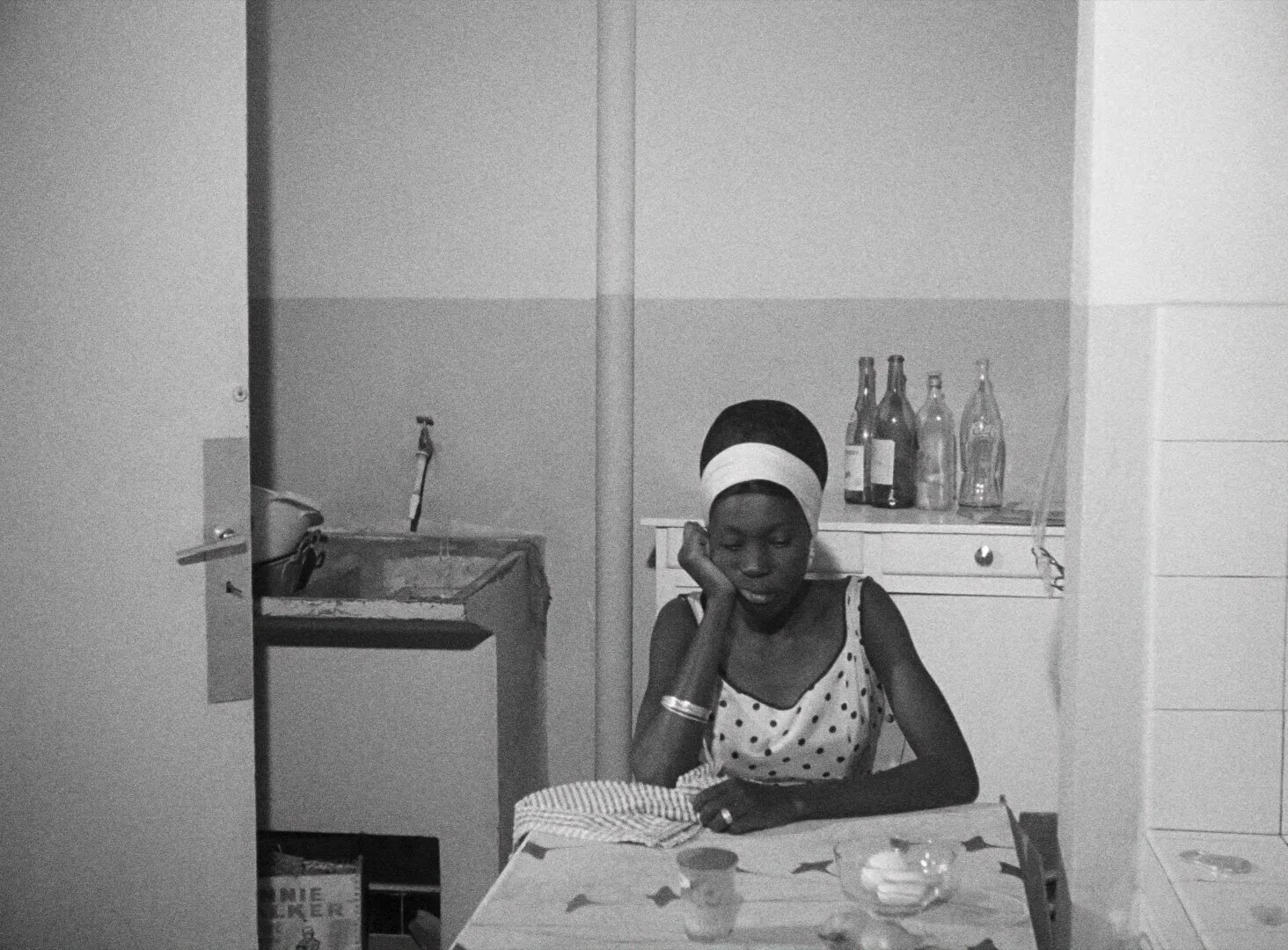 Diouana seated at a kitchen table, elbow resting on the surface, looking downward in quiet resignation — a still from Black Girl (La Noire de..., 1966) by Ousmane Sembène