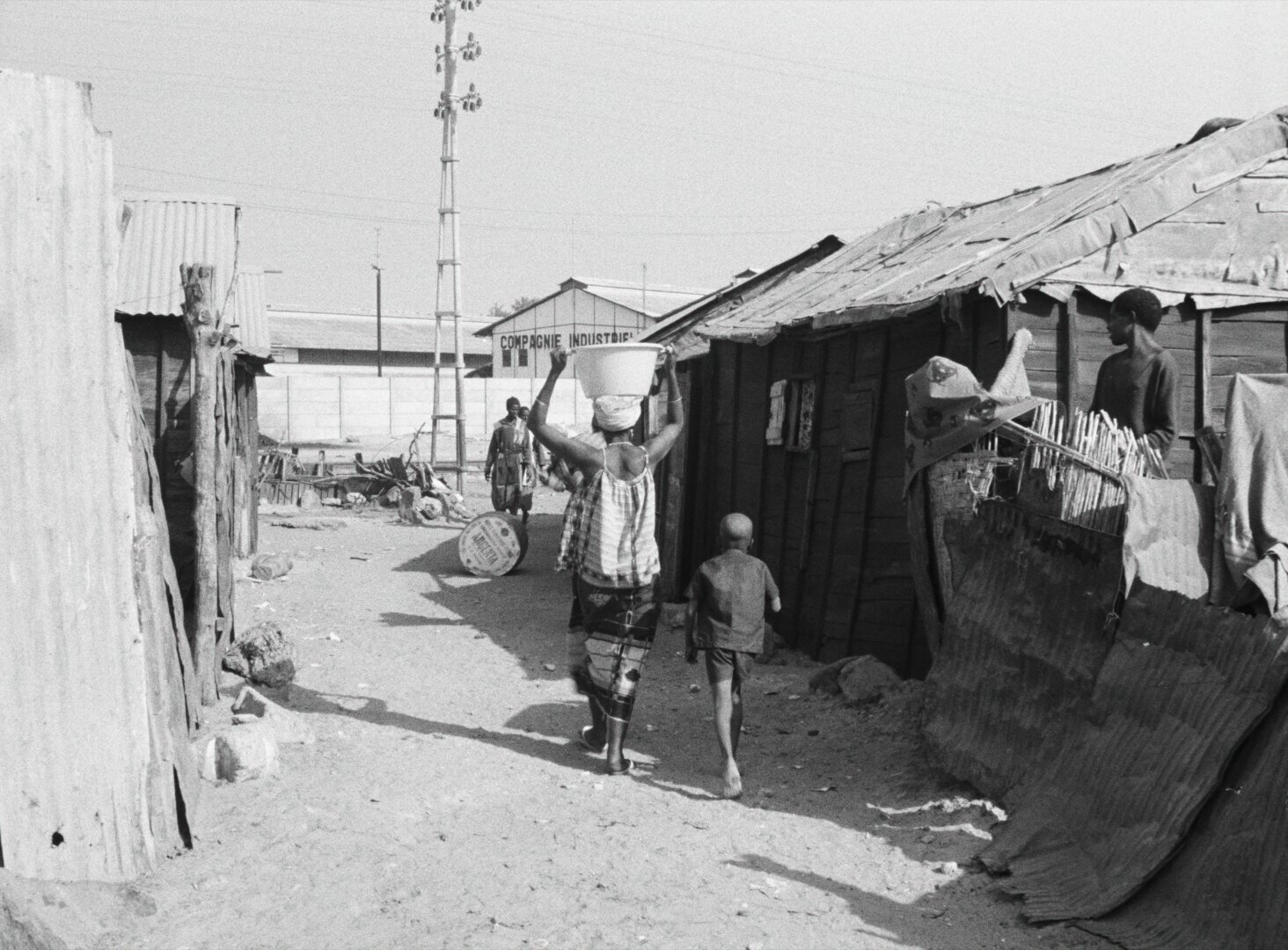 A bustling shantytown street in Dakar, a woman carrying a bucket on her head, corrugated iron rooftops under open sky — a still from Black Girl (La Noire de..., 1966) by Ousmane Sembène