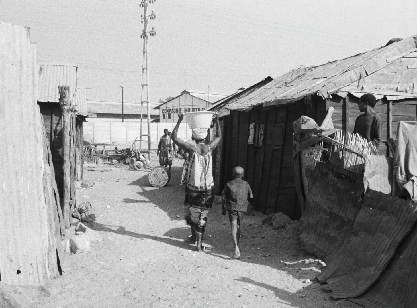 A bustling shantytown street in Dakar, a woman carrying a bucket on her head, corrugated iron rooftops under open sky — a still from Black Girl (La Noire de..., 1966) by Ousmane Sembène