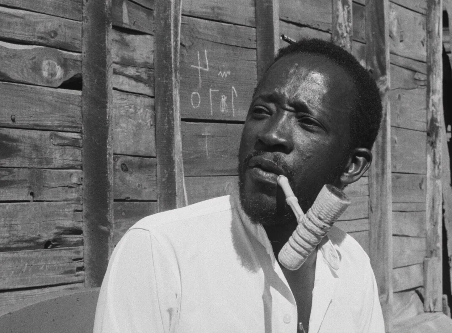 A man smokes a pipe outside a weathered wooden wall in Dakar — a still from Black Girl (La Noire de..., 1966) by Ousmane Sembène