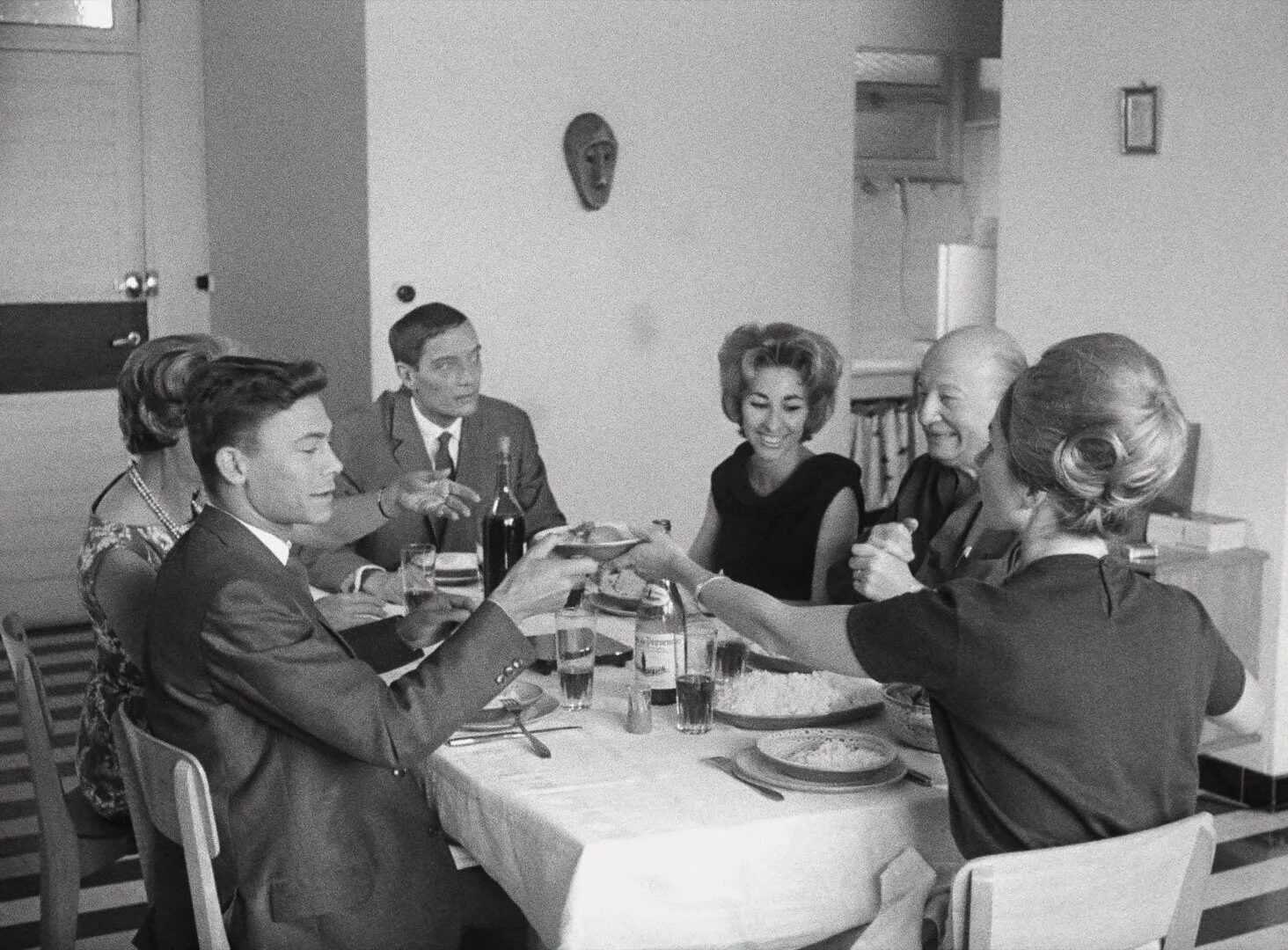 A French family gathered around a dinner table, animated in conversation, an African mask visible on the wall behind them — a still from Black Girl (La Noire de..., 1966) by Ousmane Sembène