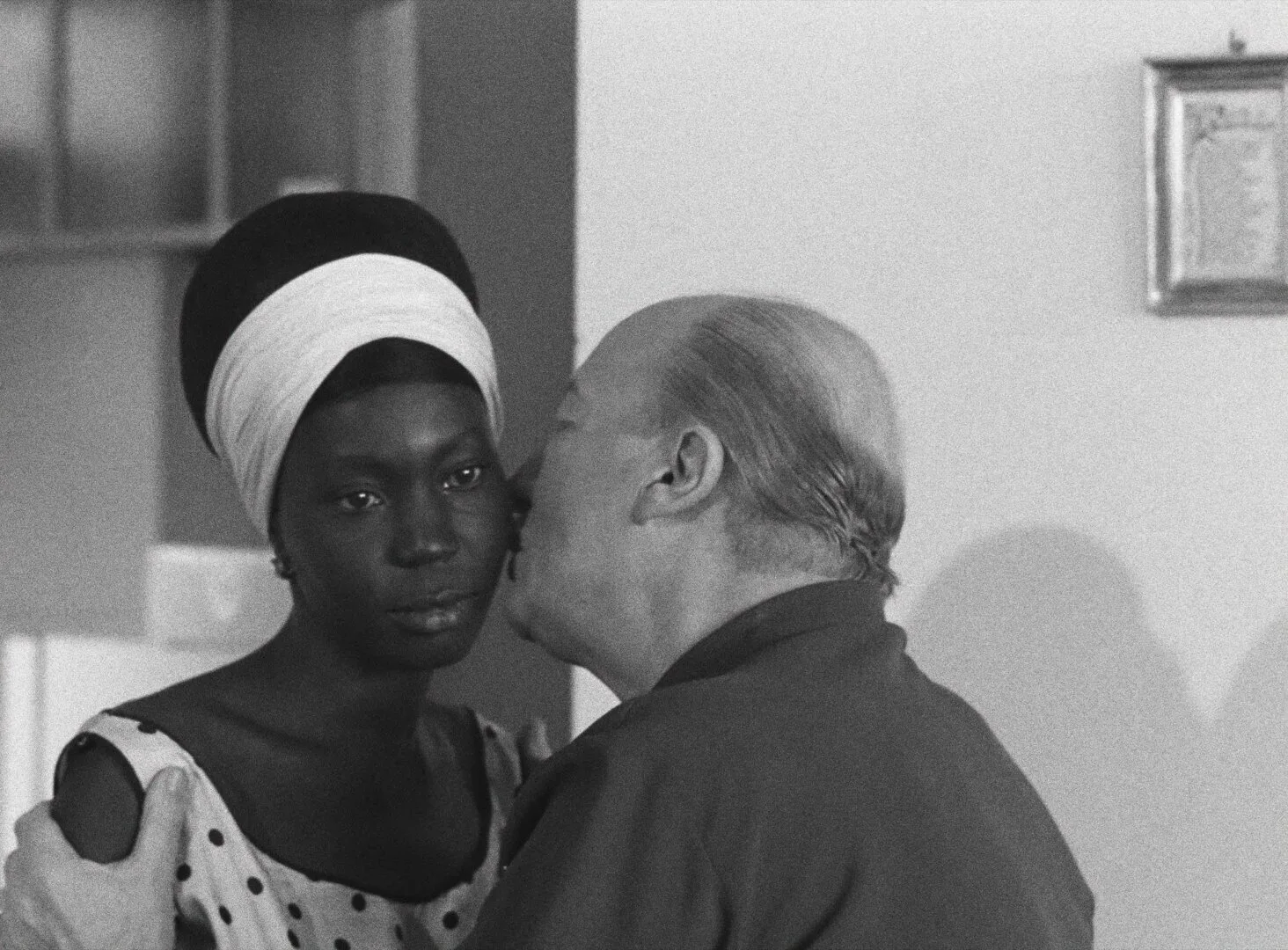 An older French man kisses Diouana on the cheek as she stands still, her expression distant and unreadable — a still from Black Girl (La Noire de..., 1966) by Ousmane Sembène
