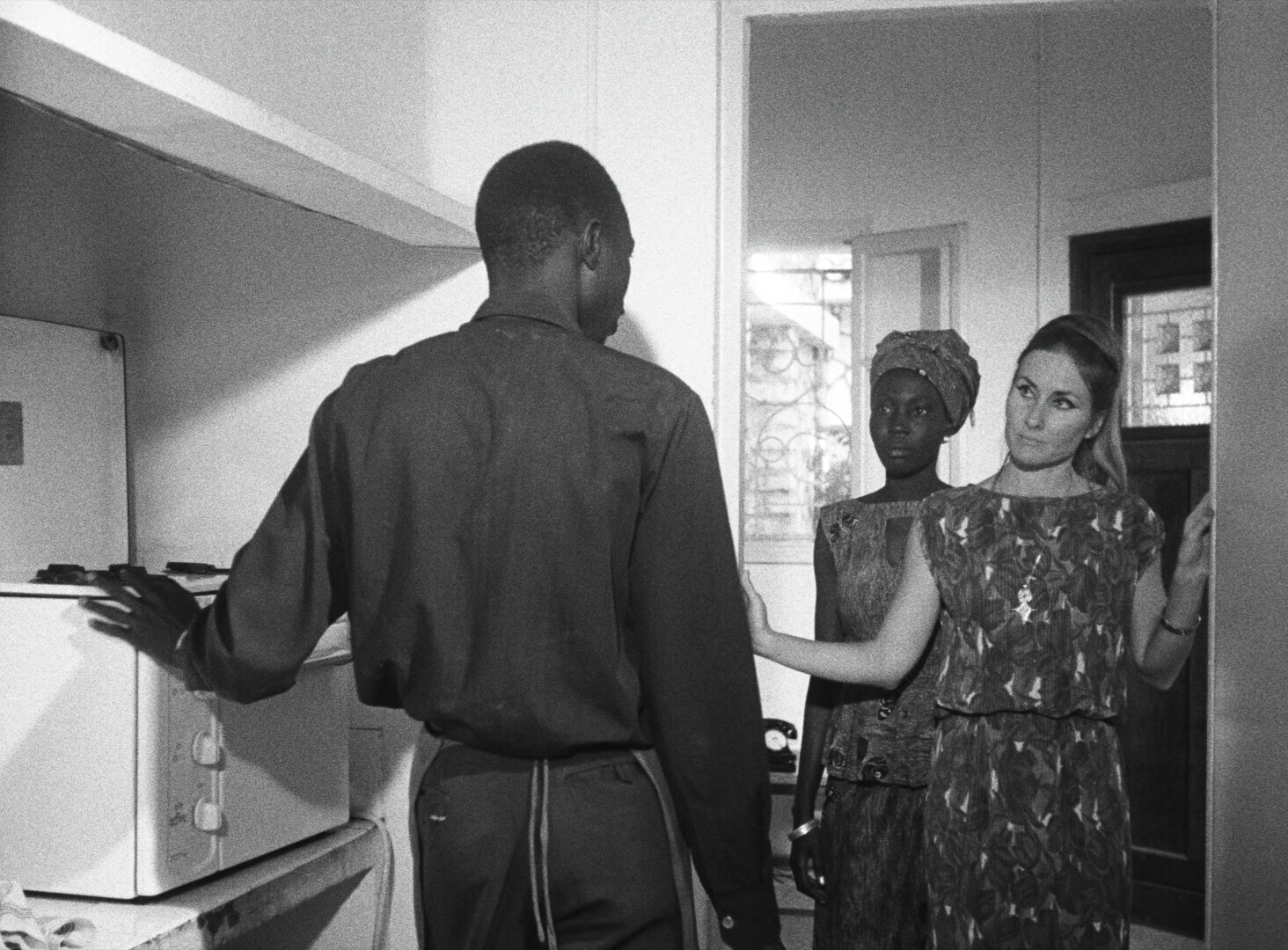 Diouana stands in the background of a kitchen doorway, half-obscured, watching as her French employer and a man speak in the foreground — a still from Black Girl (La Noire de..., 1966) by Ousmane Sembène
