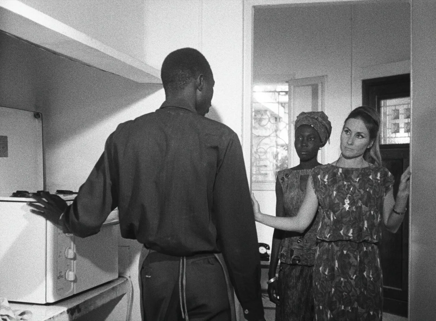 Diouana stands in the background of a kitchen doorway, half-obscured, watching as her French employer and a man speak in the foreground — a still from Black Girl (La Noire de..., 1966) by Ousmane Sembène