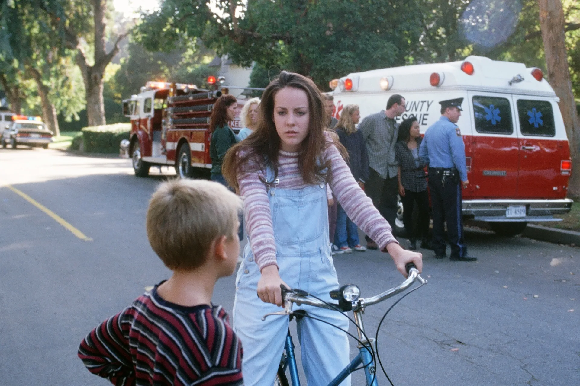 A girl on a bicycle pauses near fire trucks and an ambulance in Donnie Darko (2001)