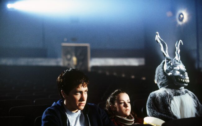 Jake Gyllenhaal as Donnie and Jena Malone as Gretchen sit in an empty cinema beside Frank the rabbit, lit by the projector beam, in Donnie Darko (2001)