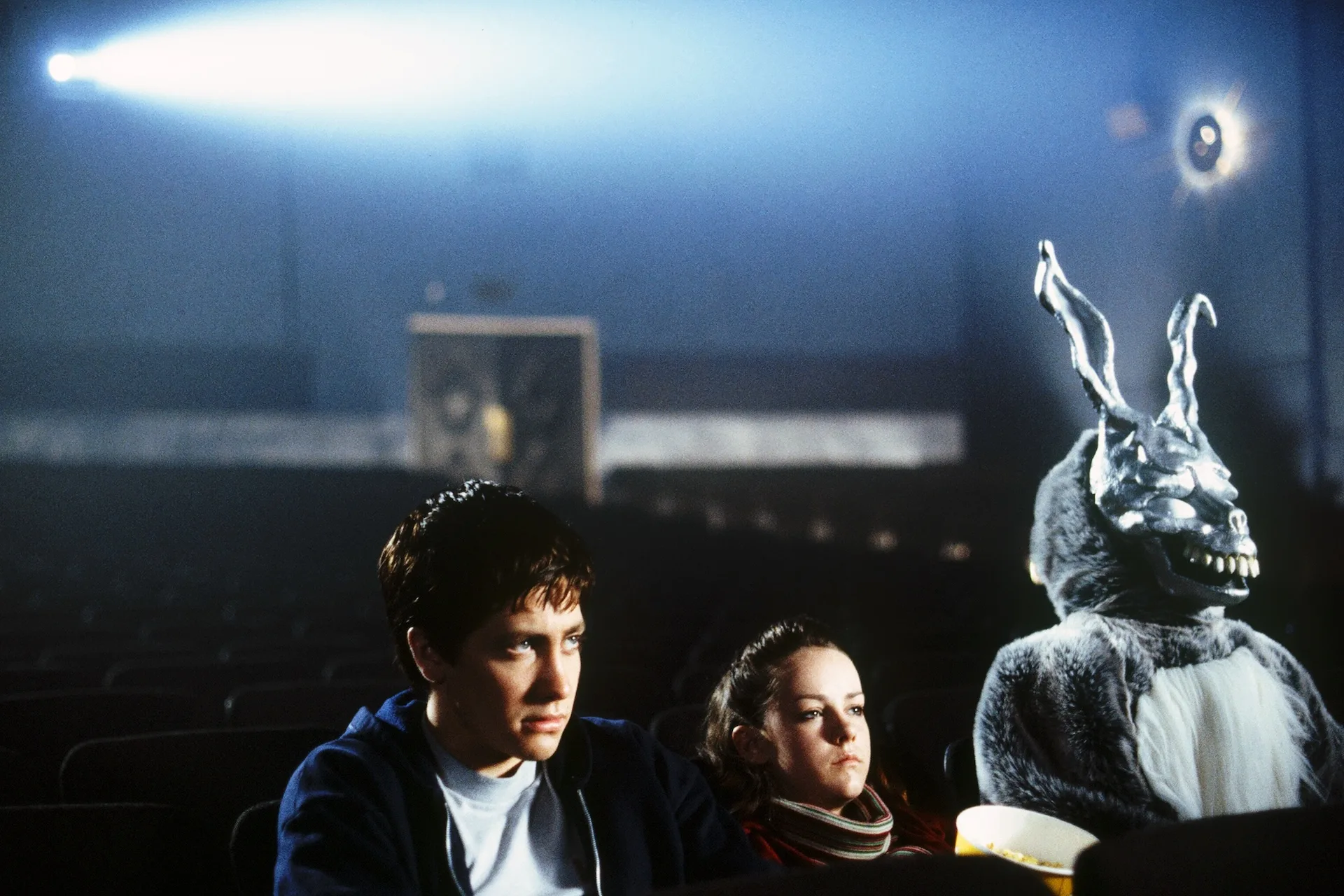 Jake Gyllenhaal as Donnie and Jena Malone as Gretchen sit in an empty cinema beside Frank the rabbit, lit by the projector beam, in Donnie Darko (2001)