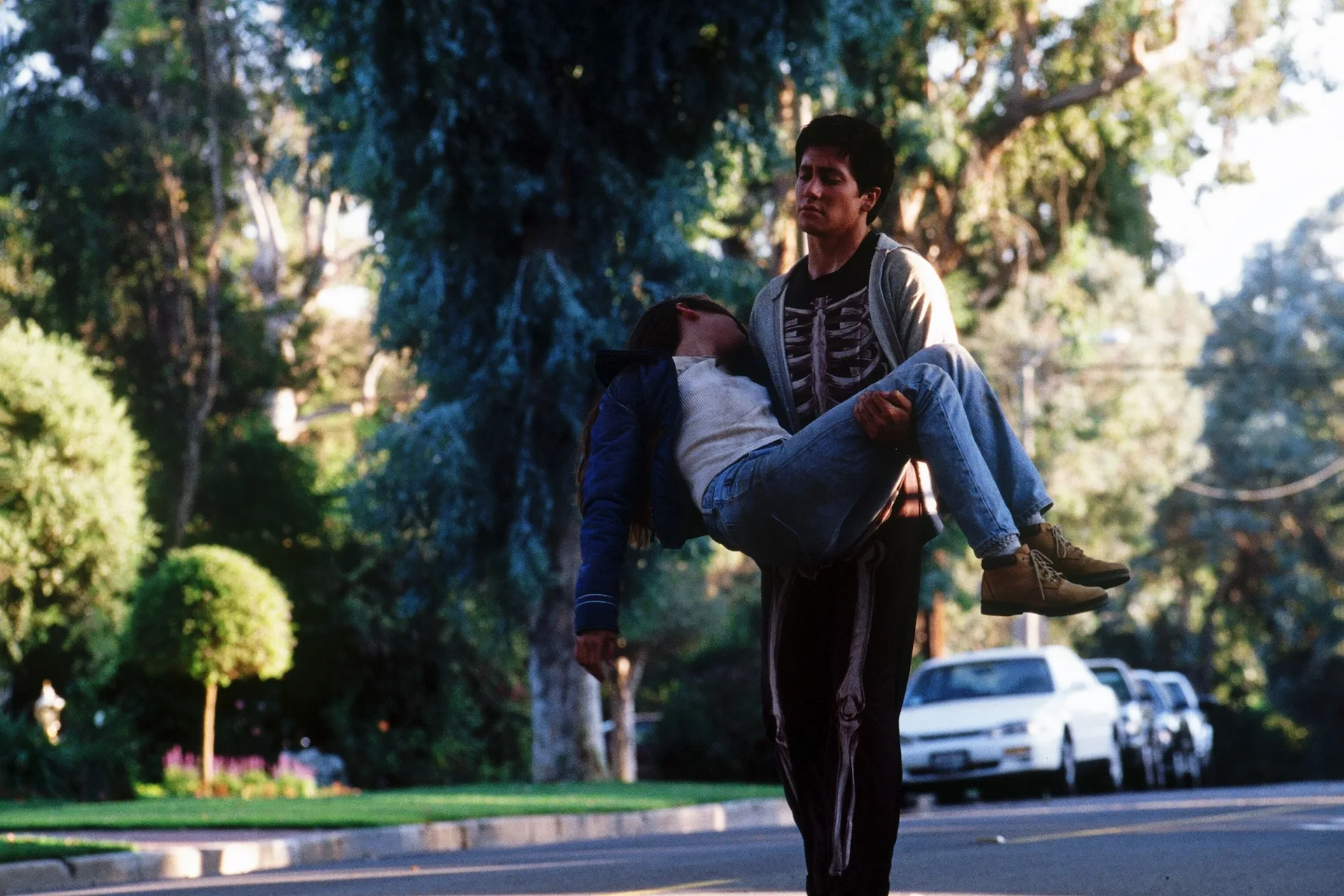 Jake Gyllenhaal as Donnie, in his skeleton costume, carries an unconscious Jena Malone as Gretchen down a tree-lined street in Donnie Darko (2001)