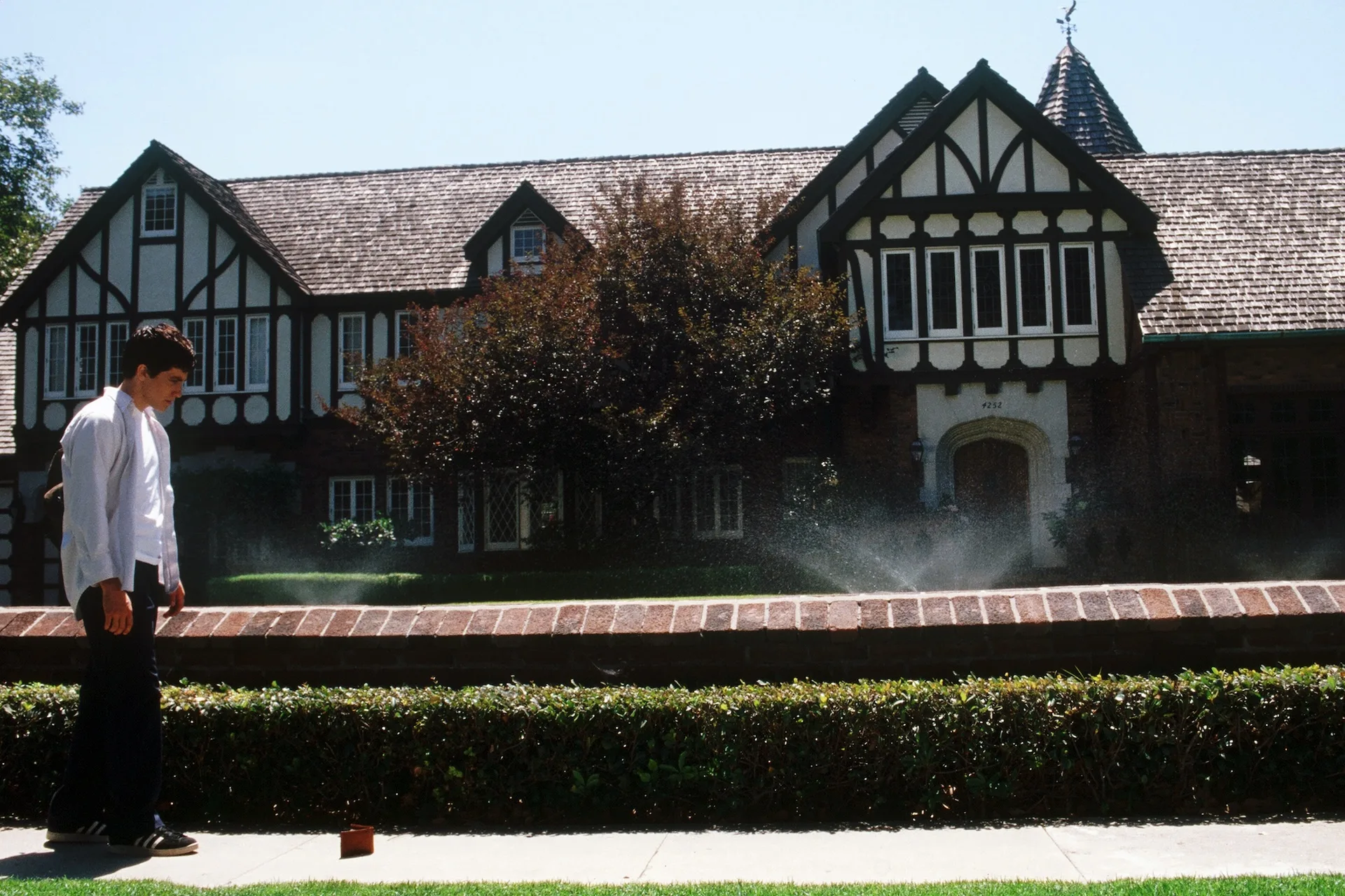 Jake Gyllenhaal as Donnie walks past a Tudor suburban house as the sprinklers run in Donnie Darko (2001)