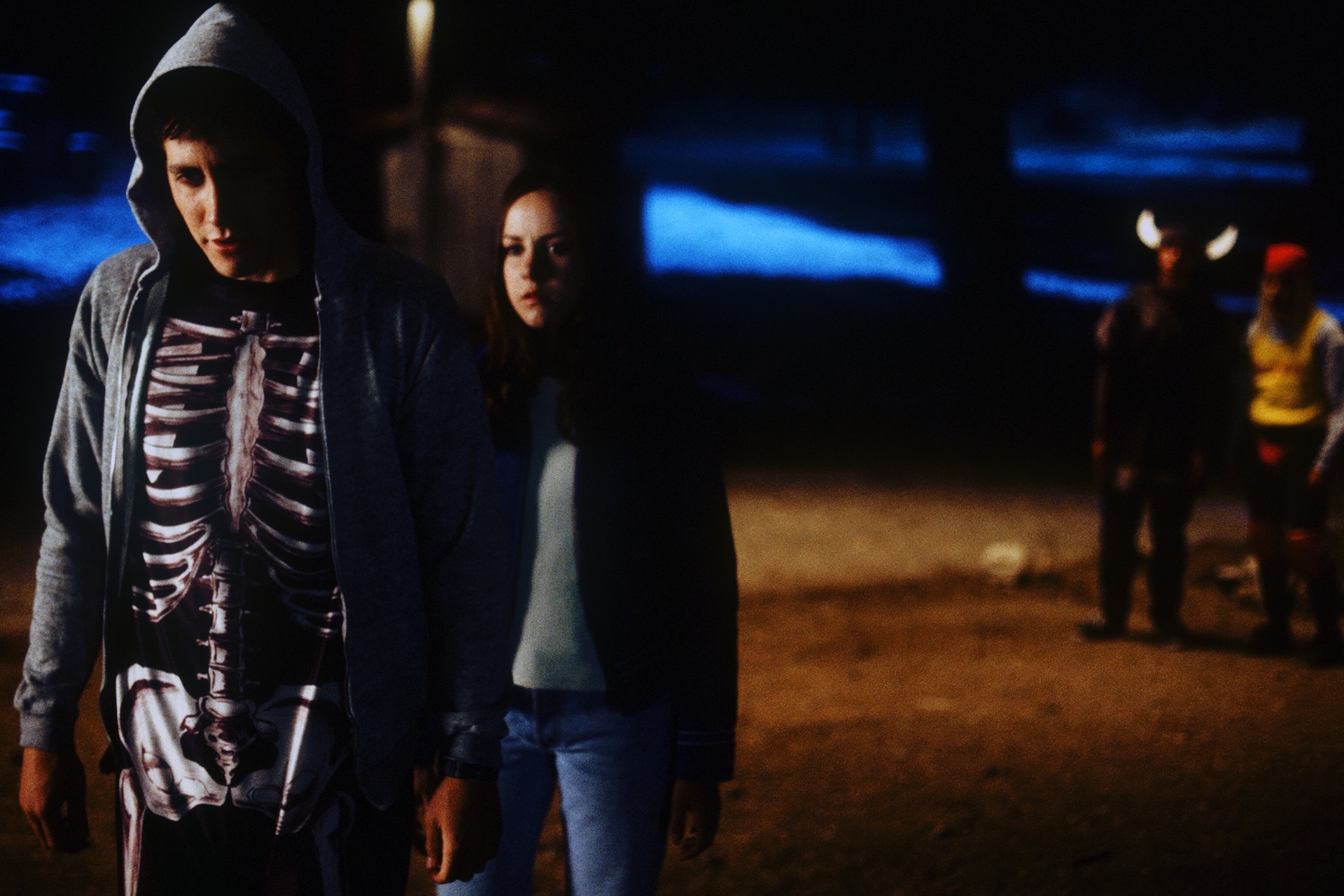Jake Gyllenhaal as Donnie, in his skeleton costume, walks with Jena Malone as Gretchen past costumed figures on Halloween night in Donnie Darko (2001)