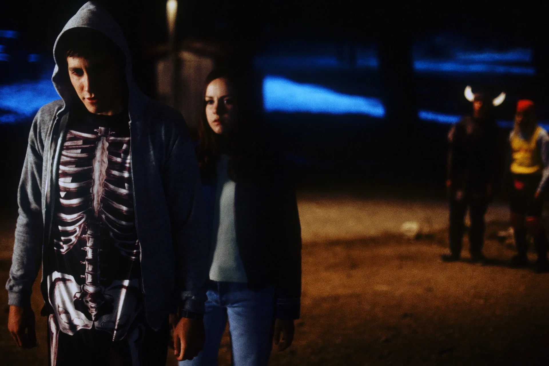 Jake Gyllenhaal as Donnie, in his skeleton costume, walks with Jena Malone as Gretchen past costumed figures on Halloween night in Donnie Darko (2001)