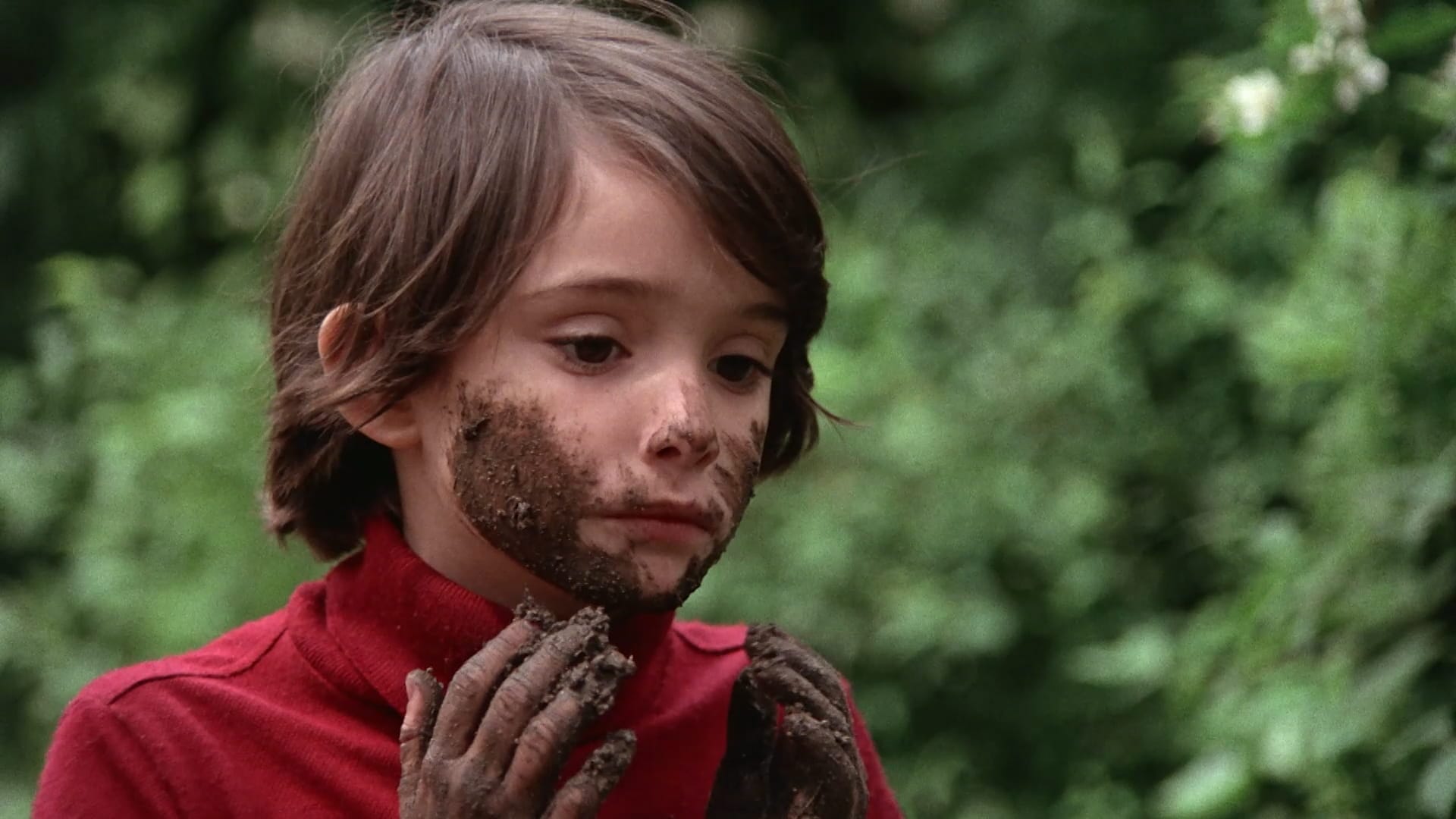 Ana looks at her mud-covered hands, face smeared with dirt, against garden greenery in Cría Cuervos