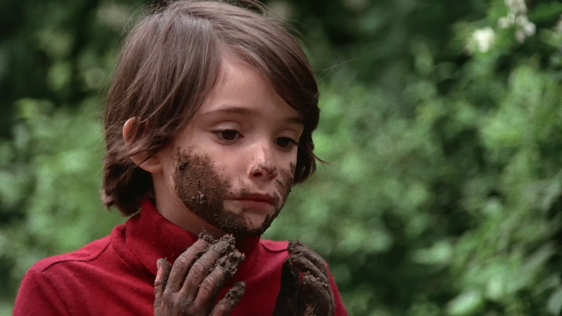 Ana looks at her mud-covered hands, face smeared with dirt, against garden greenery in Cría Cuervos