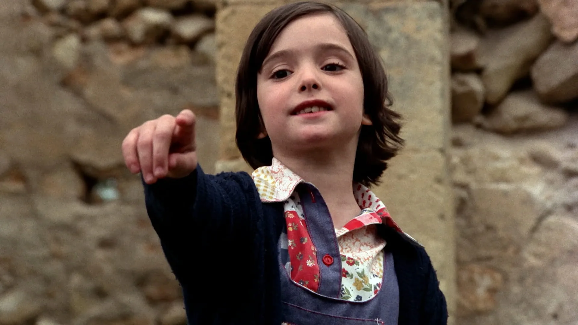 Ana points at something off-screen while playing against a stone wall in Cría Cuervos