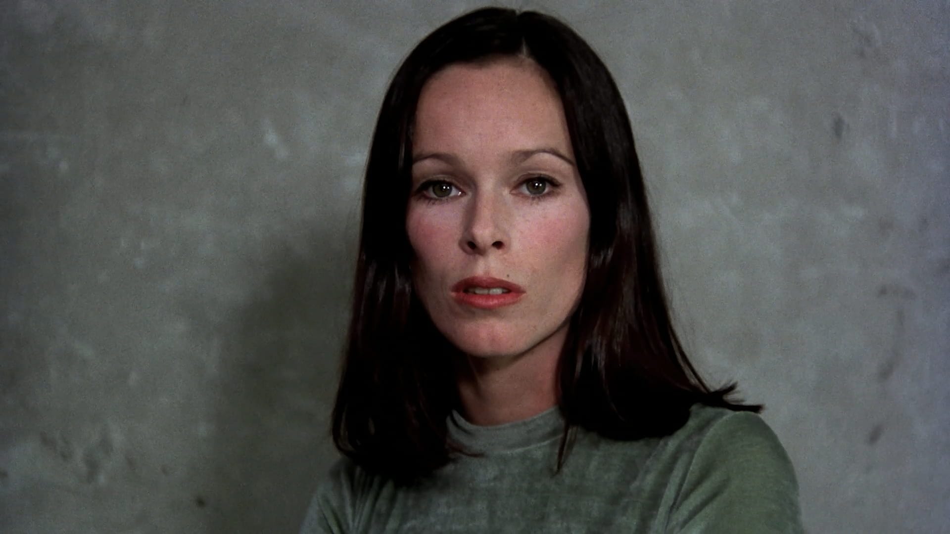 Geraldine Chaplin as the adult Ana speaking directly to camera against a grey wall in Cría Cuervos