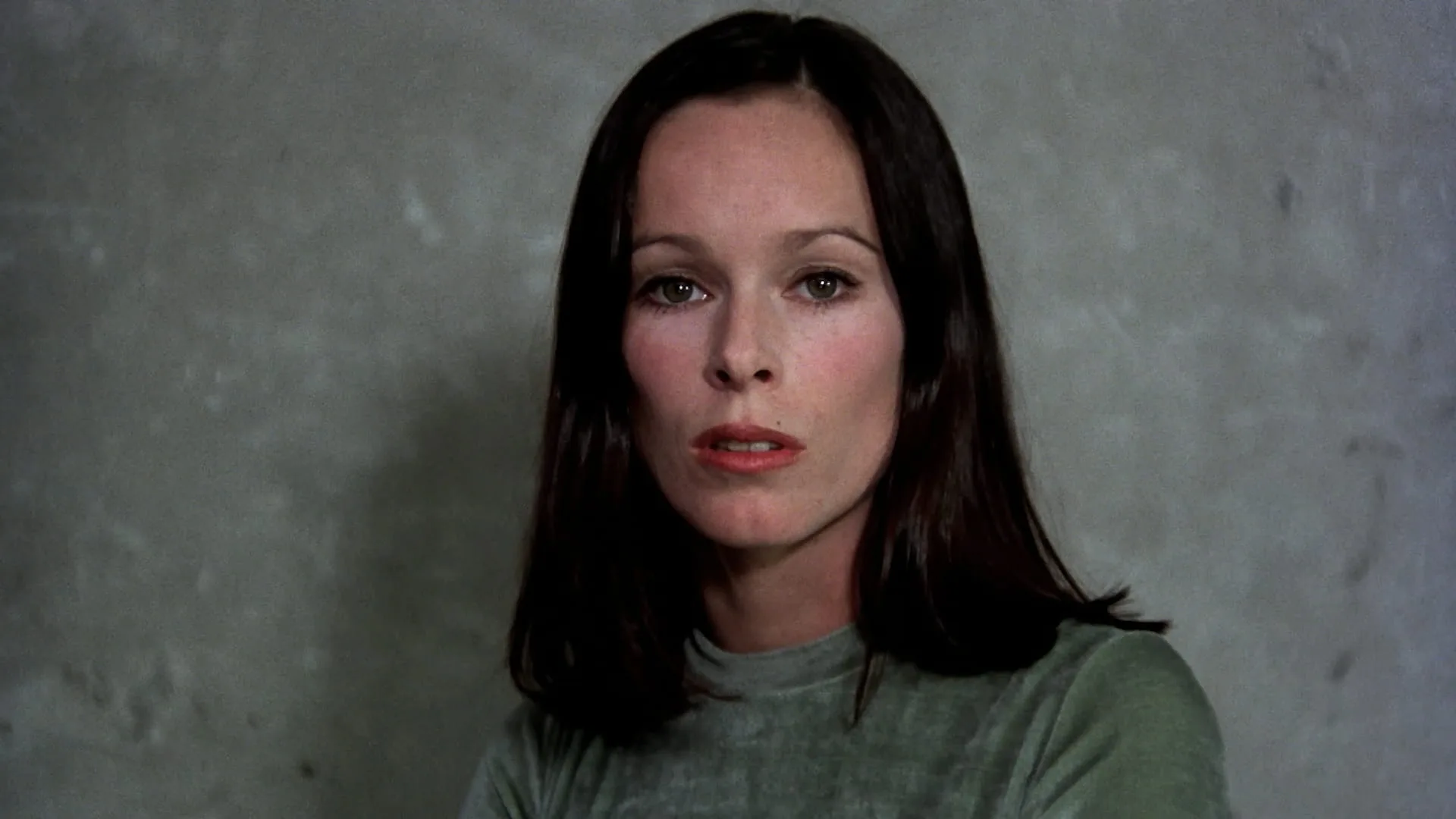 Geraldine Chaplin as the adult Ana speaking directly to camera against a grey wall in Cría Cuervos